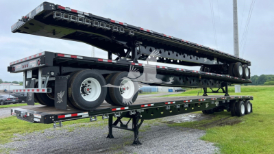 Flatbed trailer by Manac with folded deck raised, dual rear axles, and landing gear visible.