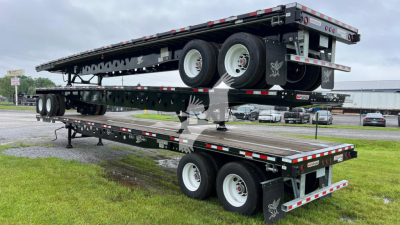 Manac steel flatbed trailer on dual-level tandems with spare tire and reflective stripes in a yard.