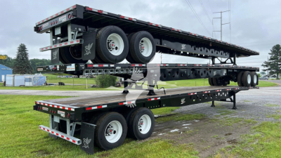 Manac steel flatbed trailer stacked on another flatbed in a yard, dual rear axles with white rims, black frame, reflective safety tape along edges.