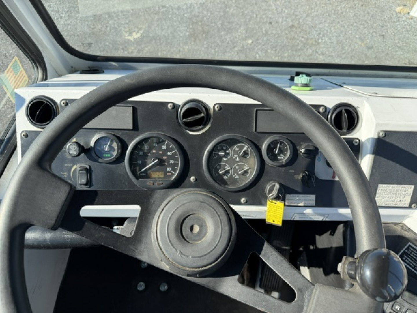 Dashboard view of a truck cab showing round gauges, steering wheel, and control switches. Visible text includes various gauge markings and a yellow tag hanging near the center console.