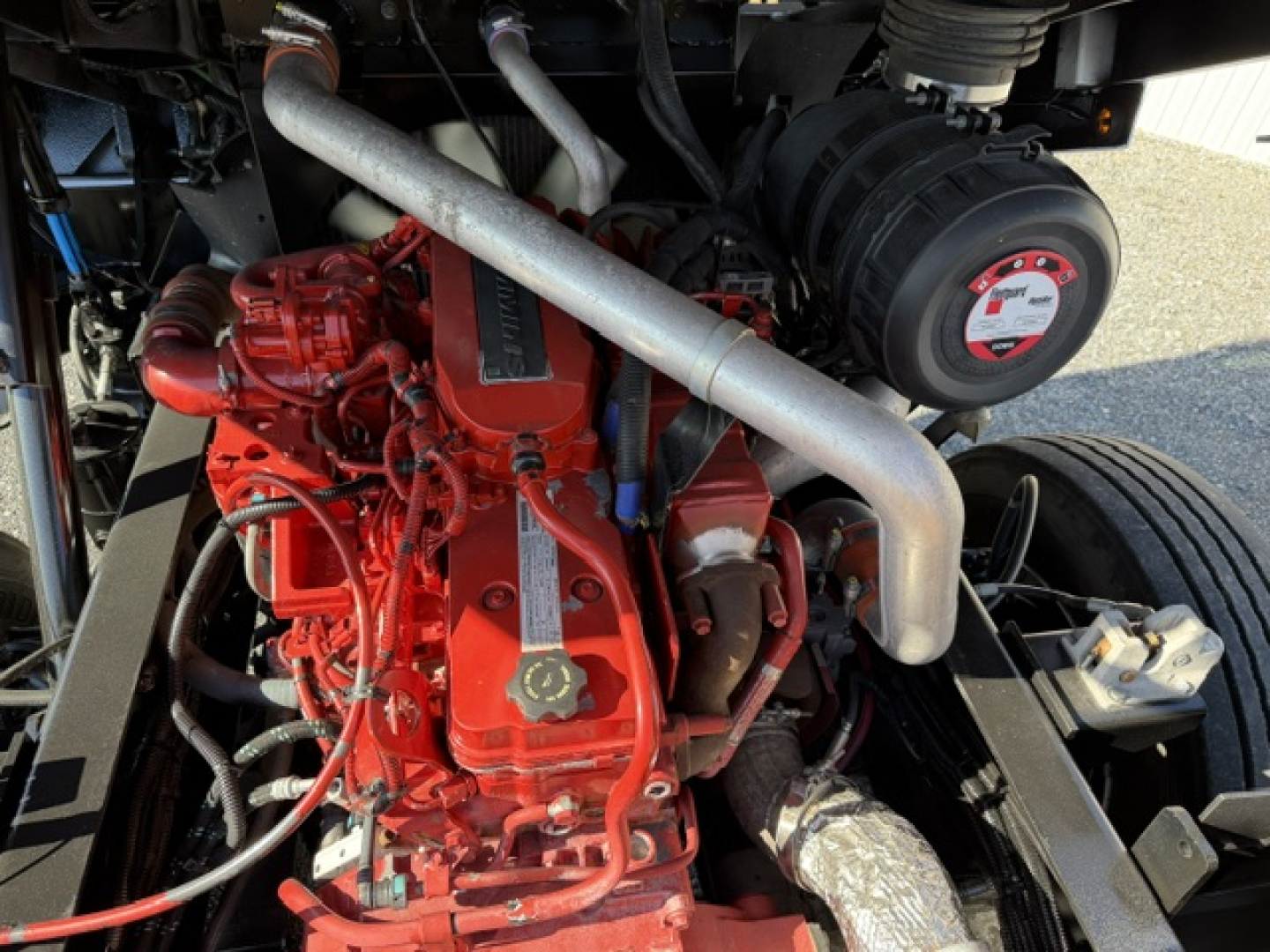 Engine bay of a 2018 Capacity Sabre DOT truck showing red engine, intake manifold, hoses, and a black circular component with a label; metallic exhaust pipe curves over the top.