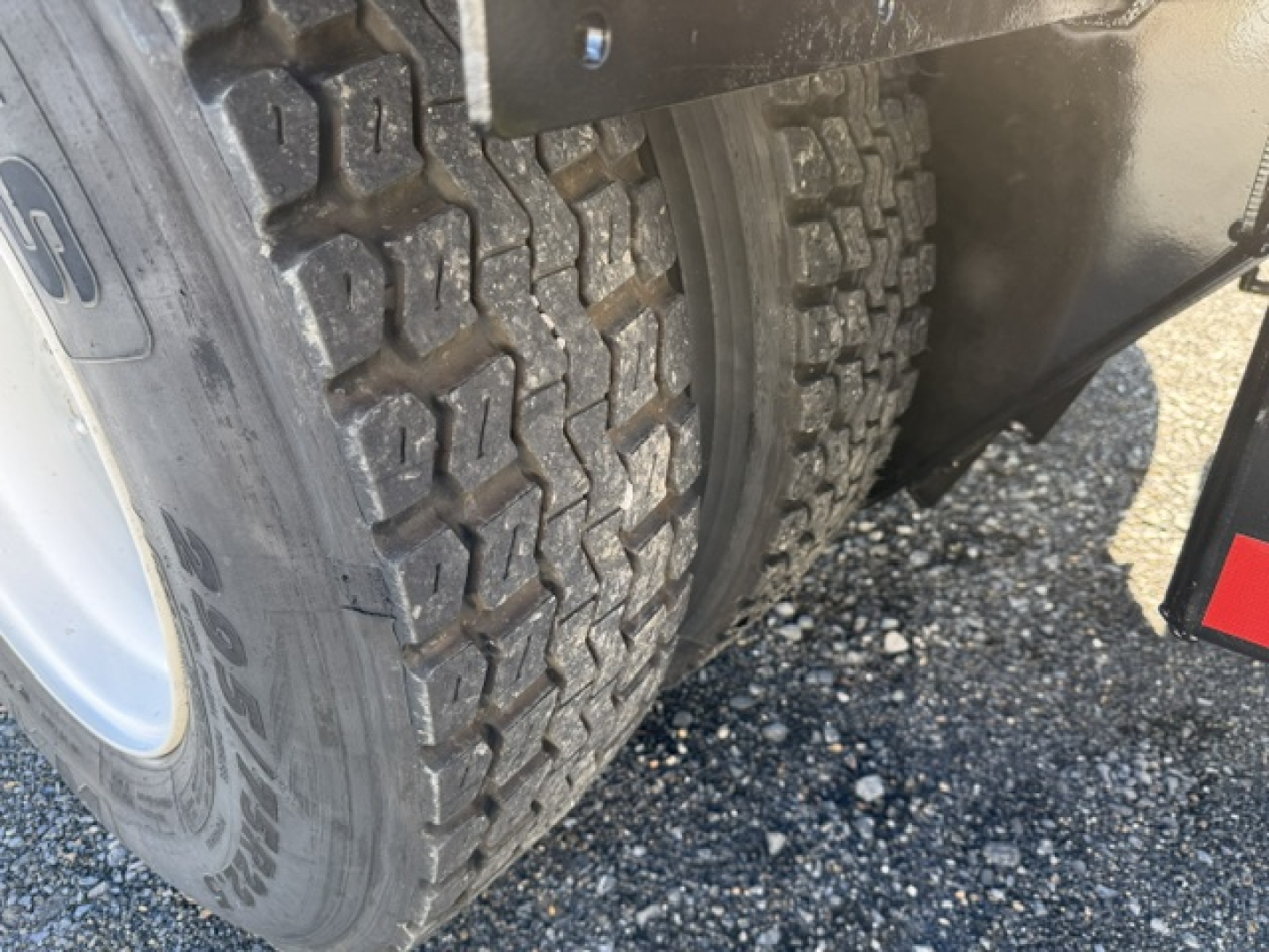 Vehicle wheel and dual rear tires showing rugged tread and part of fender; visible wheel well and gravel ground.