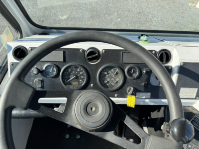 Dashboard view of a truck cab showing round gauges, steering wheel, and control switches. Visible text includes various gauge markings and a yellow tag hanging near the center console.