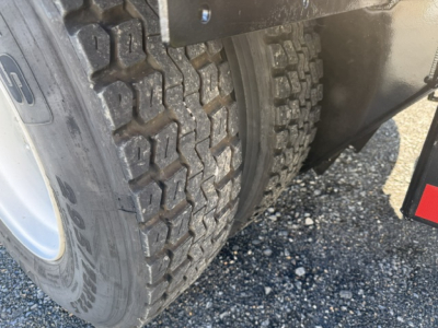 Vehicle wheel and dual rear tires showing rugged tread and part of fender; visible wheel well and gravel ground.