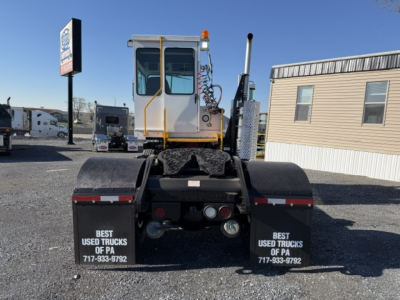 Truck rear view showing dual rear fenders with black mudflaps and warning text, cab with white paint and orange railing, exhaust stack, and rear lights.