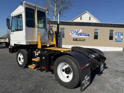 Truck chassis and cab of a yard truck with white cab, black chassis, and yellow handrails. Visible label on door, large rear wheel, and Best Used Trucks building in background.