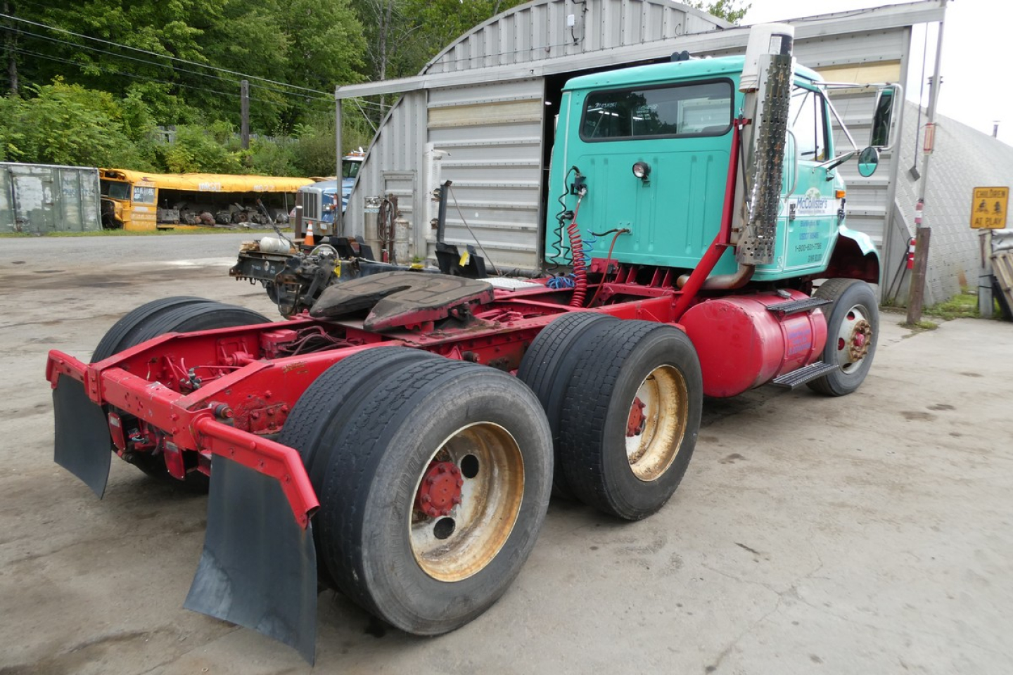 Image of a teal cab truck chassis with red frame and dual rear axles, showing the tractor’s exposed chassis and cab while parked near a building.