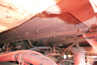 Engine bay of a truck showing hoses, cables, and components beneath the cab; view underbody with pinkish tubing and frame members.