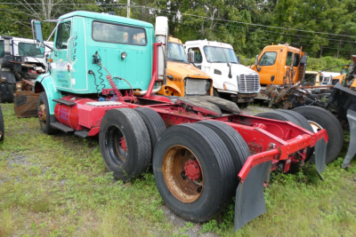 International 8100 daycab truck chassis in turquoise cab with red frame and dual rear axles, viewed from side, other orange and white trucks in background.
