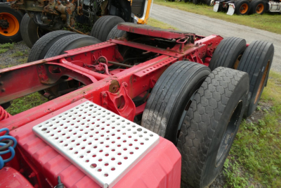 Red truck chassis with multiple rear axles and dual tires, metal perforated step plate in foreground, wheels in background.
