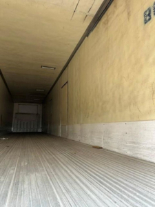 Interior of a refrigerated trailer with ribbed metal floor, yellow walls, and rear doorway. The back appears closed with a white curb at the far end.