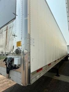 Trailer corner showing aluminum refrigerated trailer with front nose and kingpin hitch area, vertical riveted panels, and red-white reflective tape along the side.