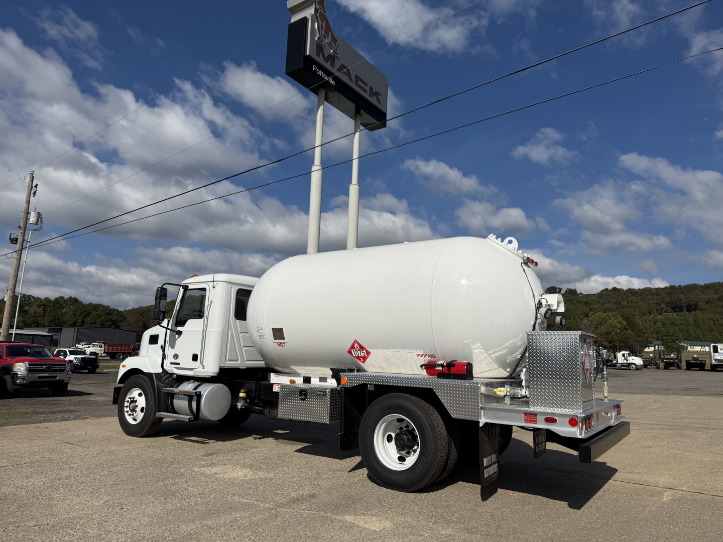 Tank truck (refueling or transport) with white cylindrical tank mounted on chassis; Mack dealership sign overhead; Diamond plate tool/storage box and rear bumper visible.