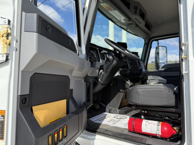 Cab interior of a 2026 Mack MD7 truck showing steering wheel, dashboard, driver seat, door pocket with a yellow card, and a red fire extinguisher on the floor mat.