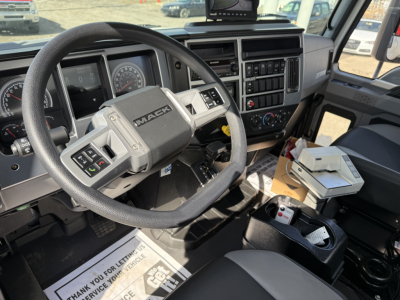 Mack MD7 truck cockpit showing steering wheel, dashboard gauges, vents, center console, and mounted device; visible paperwork and tools on the passenger side.