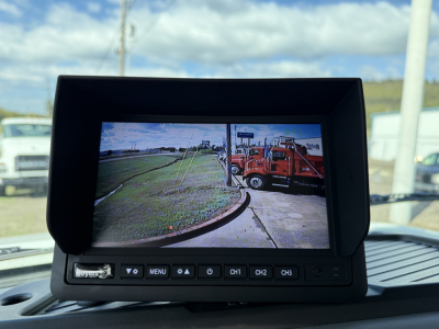 A vehicle rearview monitor display inside a Mack MD7 truck showing a side road and a red tow truck in the field of view.