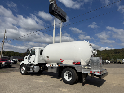 Tank truck (refueling or transport) with white cylindrical tank mounted on chassis; Mack dealership sign overhead; Diamond plate tool/storage box and rear bumper visible.
