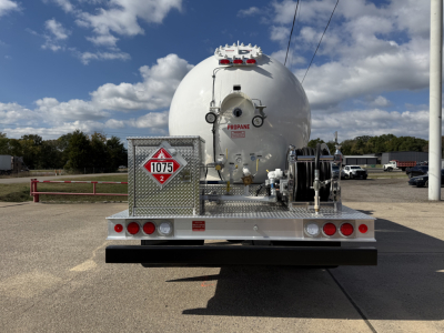 Back view of a new 2026 Mack MD7 tanker truck with a large round tank, safety placards, hoses, and rear equipment platform.