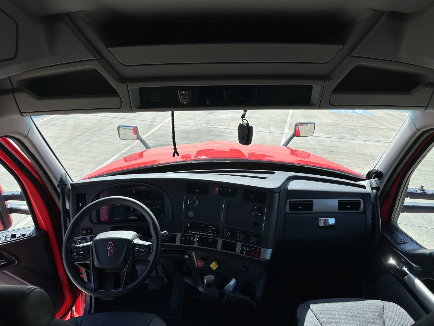 Truck cab interior of a Kenworth, steering wheel left-hand drive, dashboard with gauges, red exterior visible through windshield.