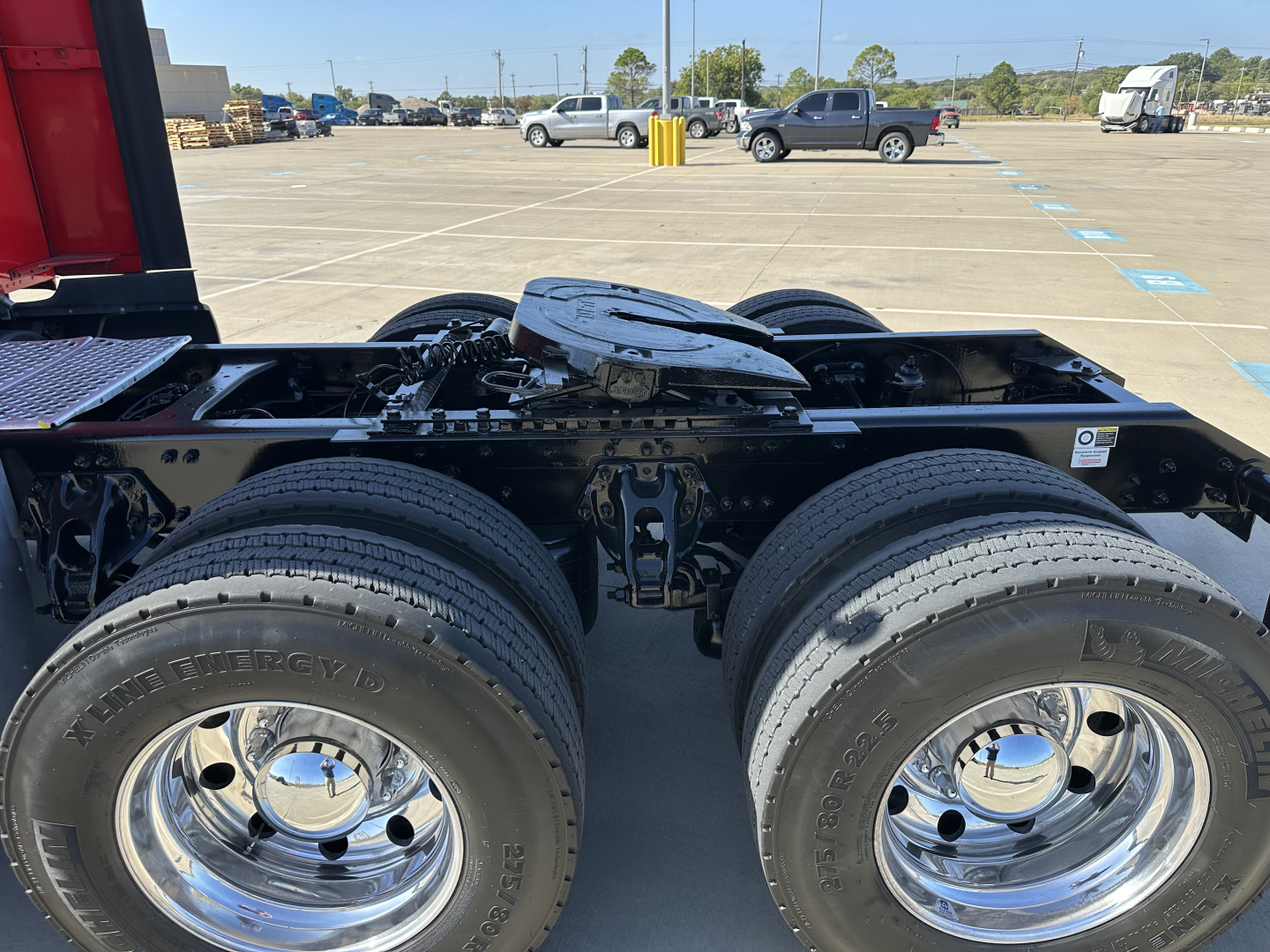 Kenworth truck chassis view showing dual rear axles and wheels with chrome rims; fuel tank area and fifth wheel visible; parking lot in background.