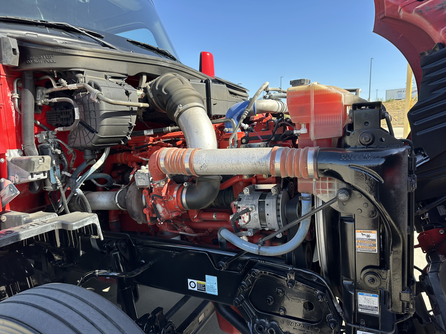 Kenworth T680 engine bay showing red engine, air intake hose, radiator coolant reservoir, and front chassis components.