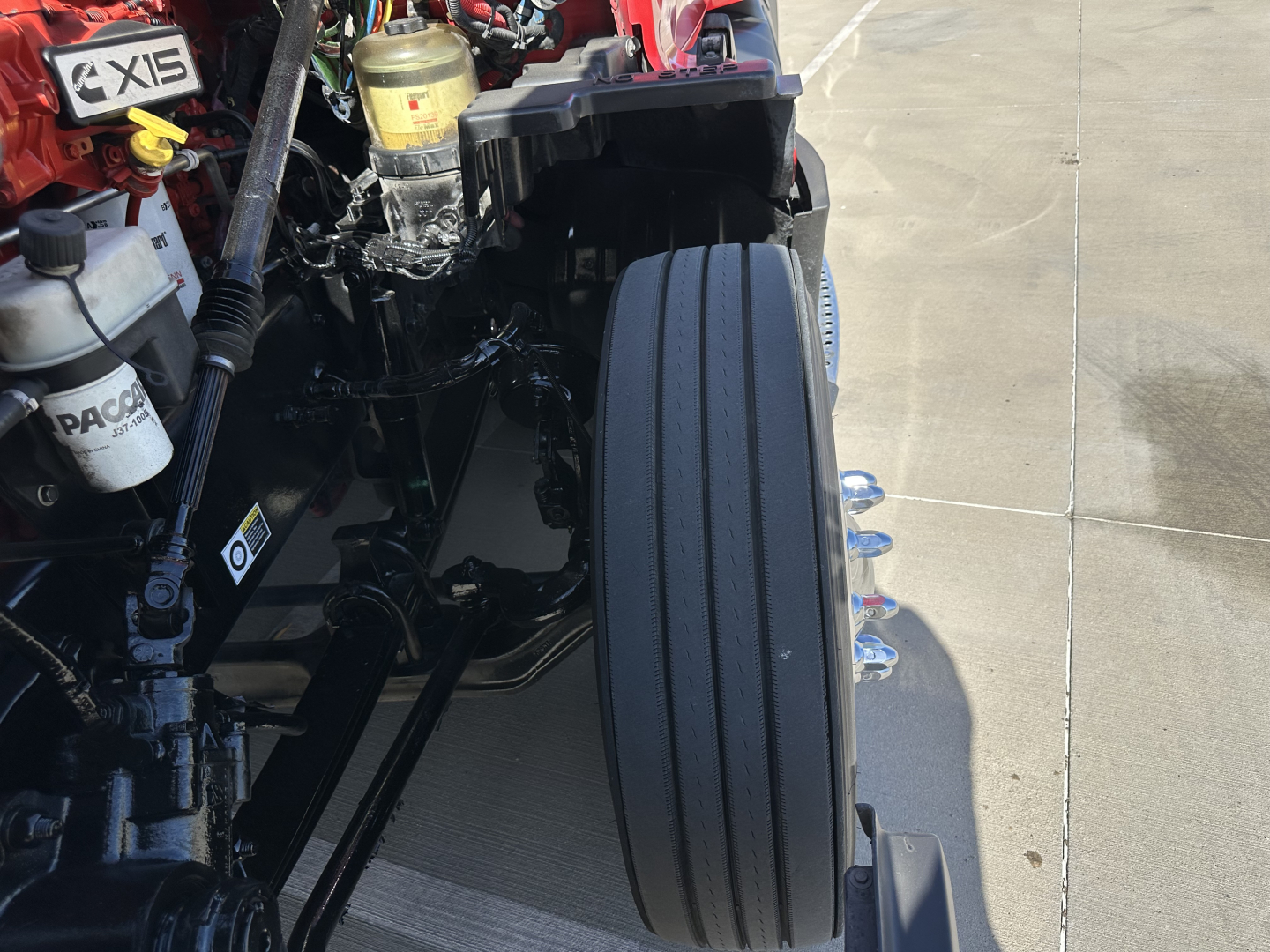 Kenworth T680 truck front end showing left front dual axle? wheel and engine bay components with hoses and reservoir caps visible.