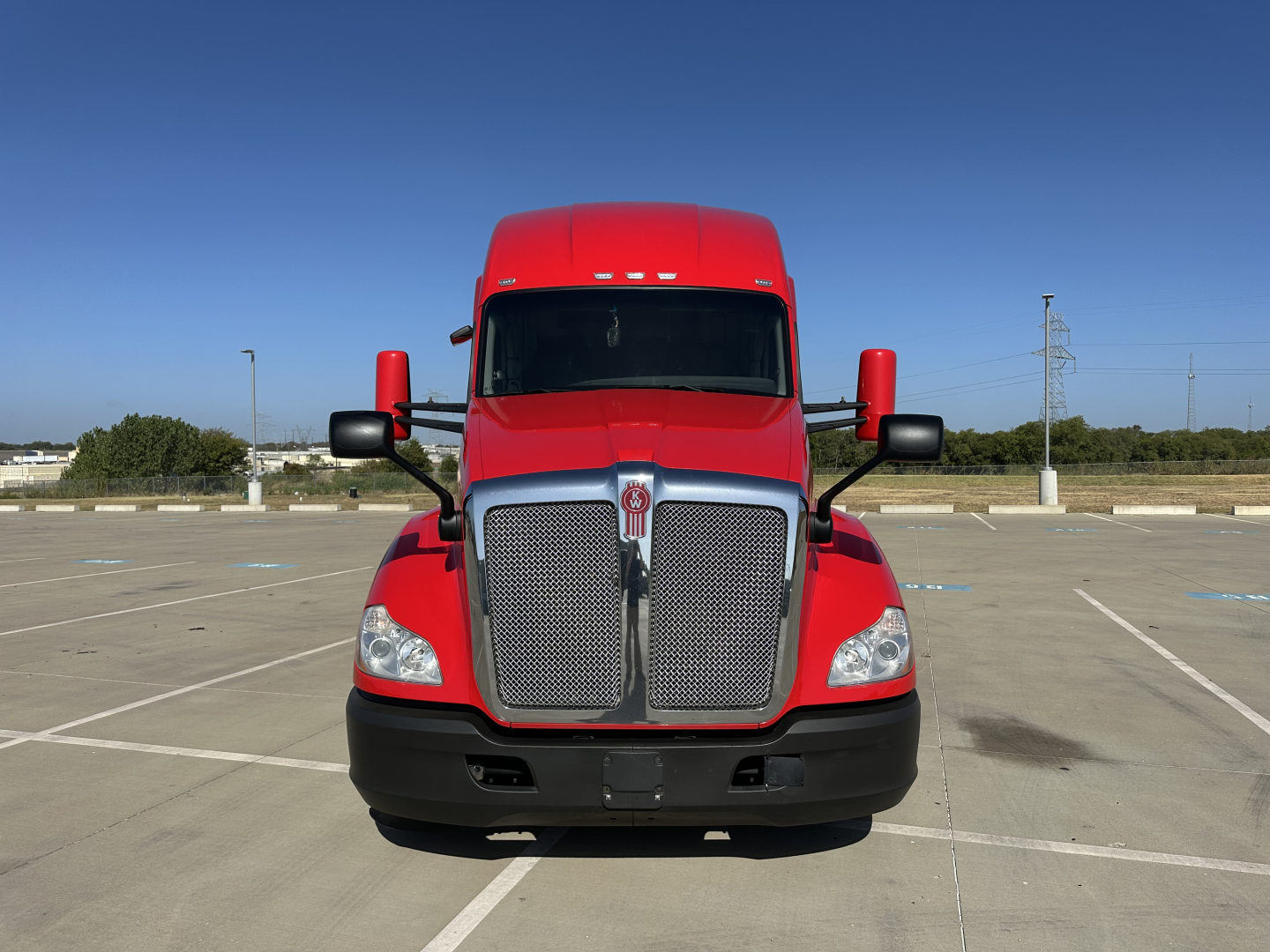 Kenworth T680 truck front view in red with chrome grille, large hood, headlights, and black bumper in an empty lot.