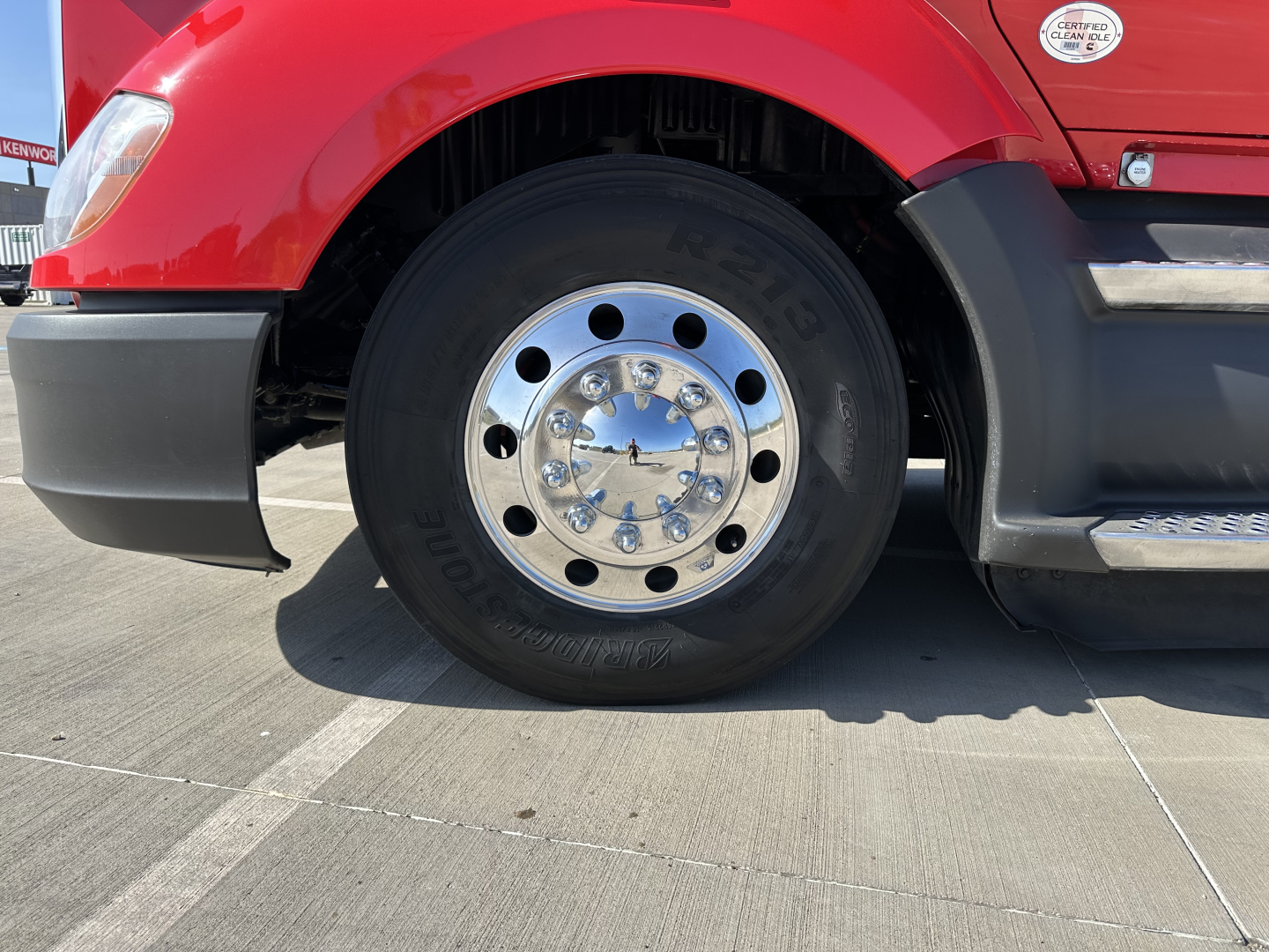 Kenworth T680 front wheel and fender assembly showing chrome hub and black tire with red cab in background.