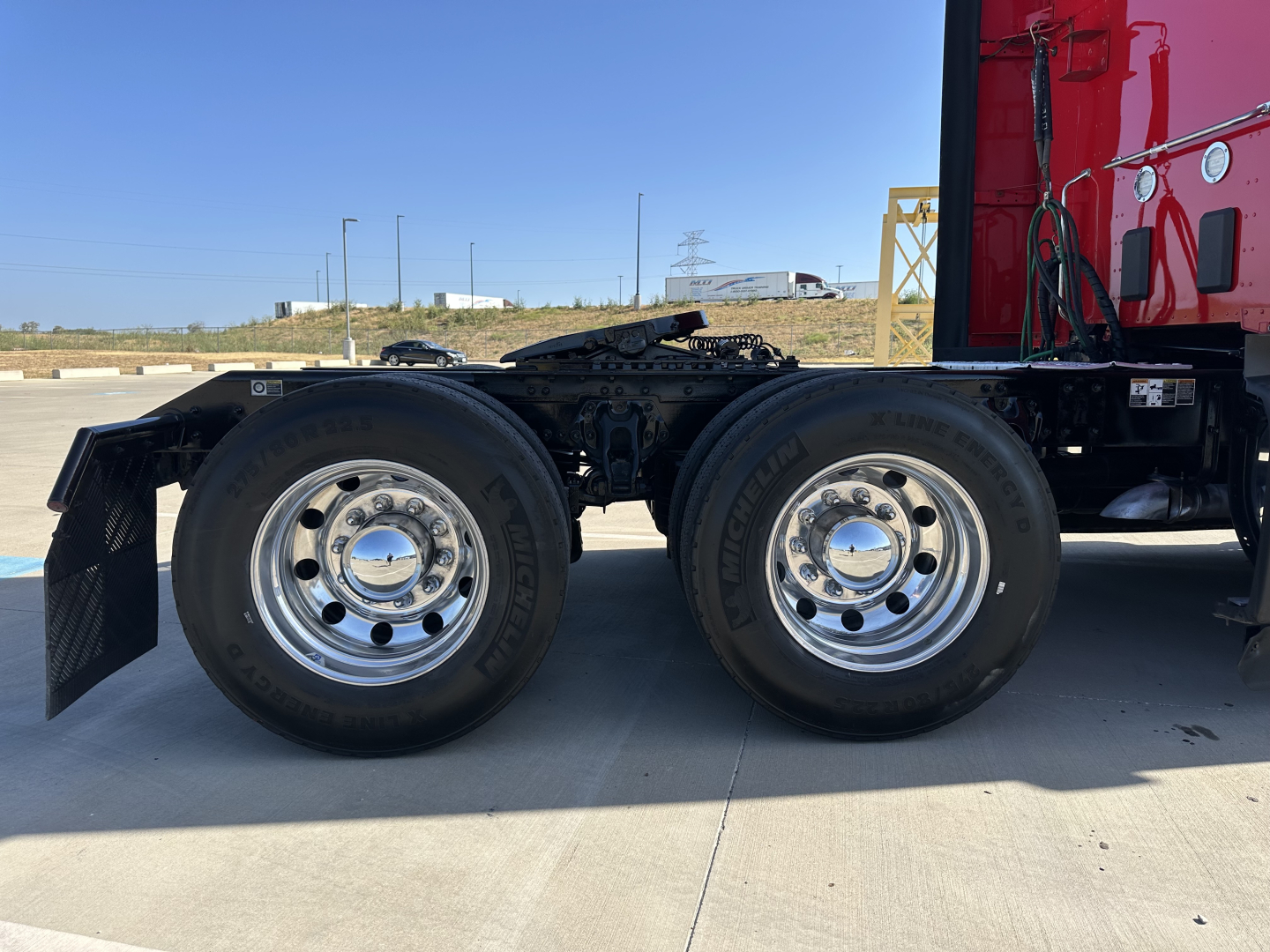 Dual rear axles of a Kenworth T680 truck chassis with chrome wheels and black mud flaps, red cab visible on right, on a sunny day.
