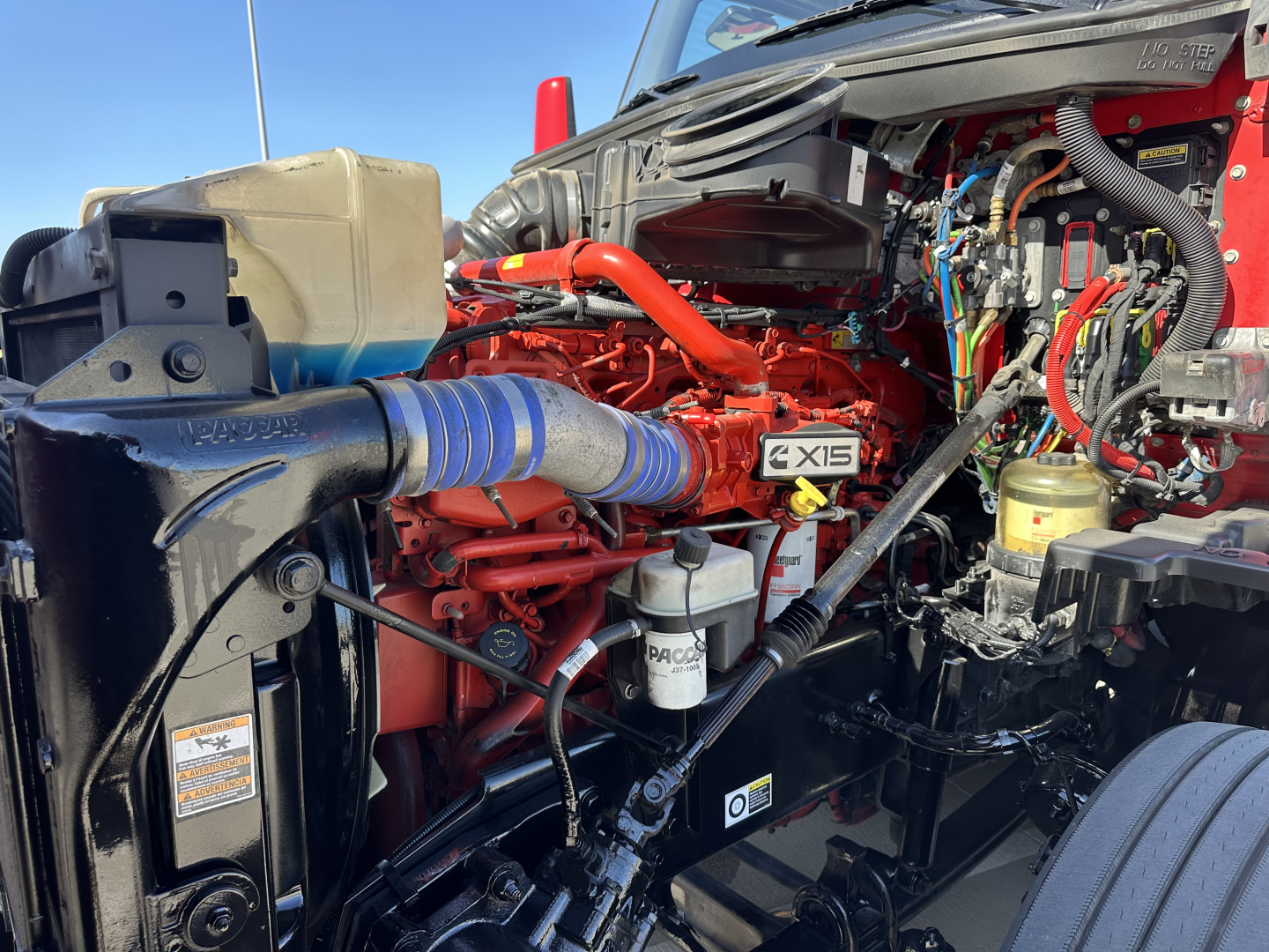 Kenworth T680 engine bay with red engine block and blue intake hose. Visible text includes X15 badge, PACCAR filter, and various hoses and wiring.