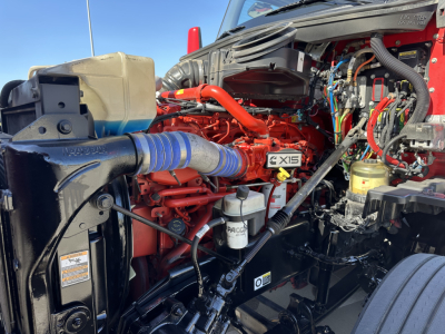 Kenworth T680 engine bay with red engine block and blue intake hose. Visible text includes X15 badge, PACCAR filter, and various hoses and wiring.