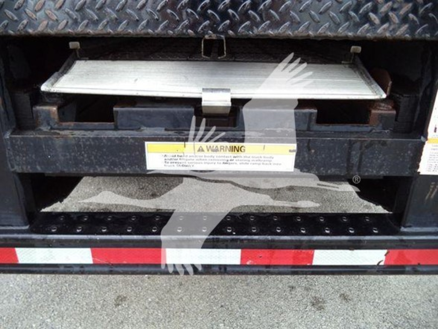 Isuzu box truck tailgate area showing underside of cargo area with warning label and metal latch, diamond plate surface above, and reflective red-white stripe on bumper.