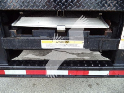 Isuzu box truck tailgate area showing underside of cargo area with warning label and metal latch, diamond plate surface above, and reflective red-white stripe on bumper.