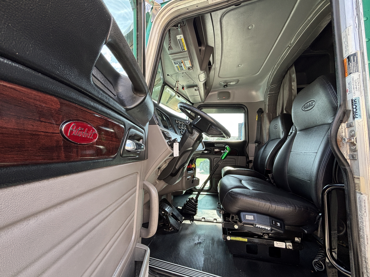 Peterbilt 389 truck cab interior view showing driver seat with black upholstery, dashboard, steering wheel, and woodgrain trim; door emblem visible.
