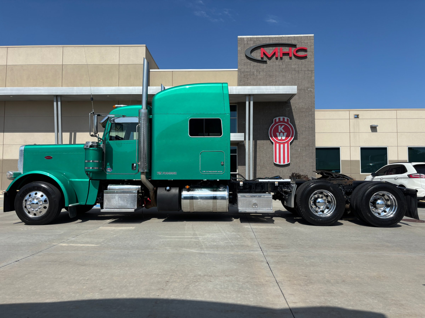 Peterbilt 389 conventional sleeper semi-truck in teal/green parked in front of a dealership building.