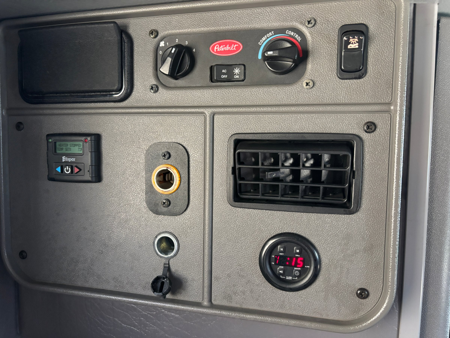 Peterbilt 389 dashboard panel showing climate controls, gauge, 12V outlet, air vent, and various switches in a sleeper truck interior.