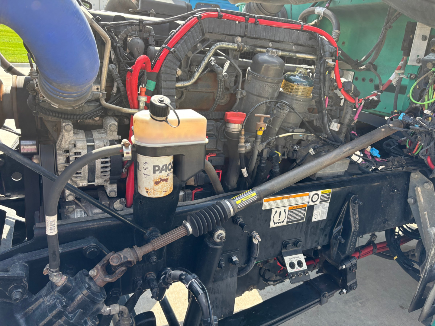 Peterbilt 389 engine bay showing coolant reservoir, hoses, and various components; visible lines, cables, and a yellowish coolant tank cap.