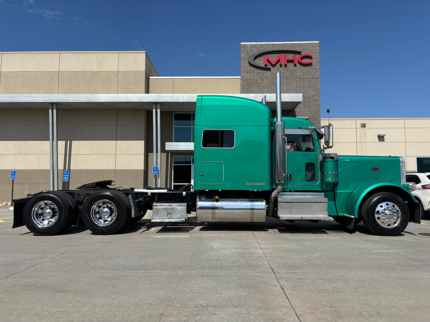 Peterbilt 389 truck side view showing green sleeper cab and chassis with dual rear axles in a dealership lot.