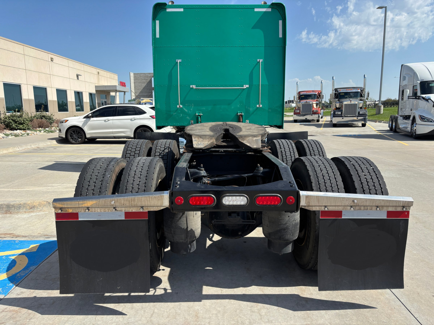 Peterbilt 389 tractor rear view showing dual rear axles, fifth wheel, rear mud flaps, and fuel/air lines. Green sleeper cab in distance, lot with other trucks.