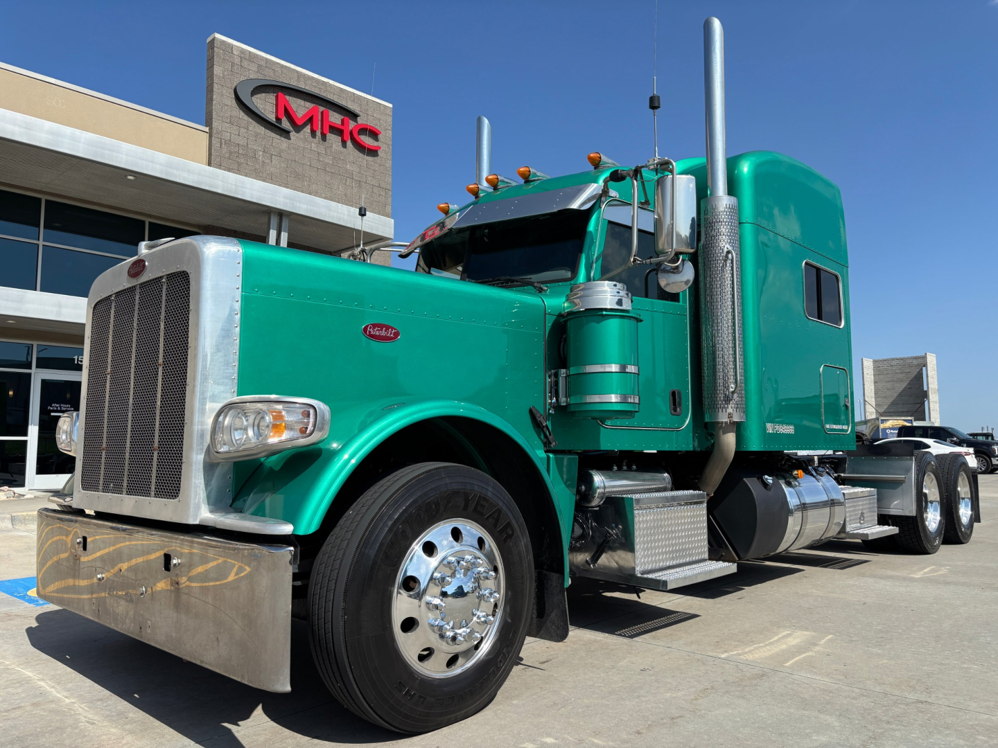 Peterbilt 389 semi truck in turquoise/green color, front grille, large chrome bumper, sleeper cab, chrome exhaust stack, parked in lot.