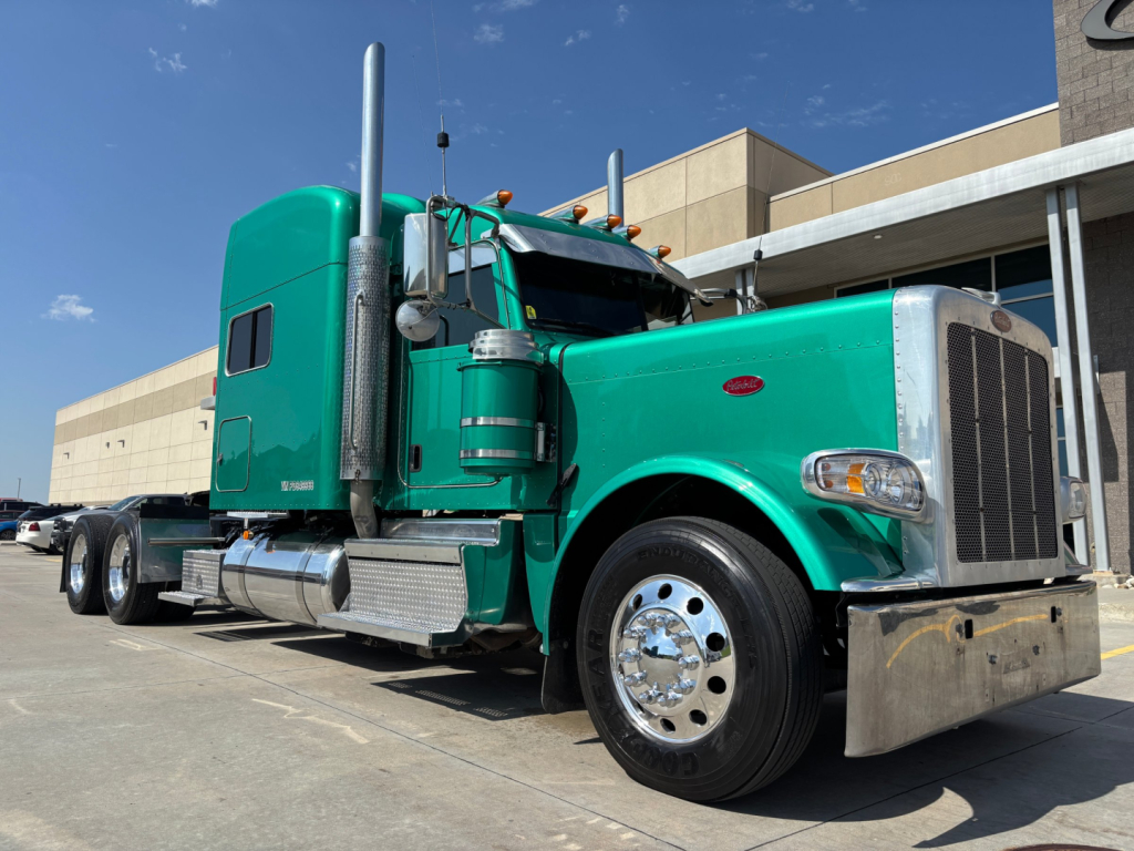 Green Peterbilt 389 diesel truck in a parking lot with sleeper cab and chrome exhaust stacks, front grille, and large bumper.