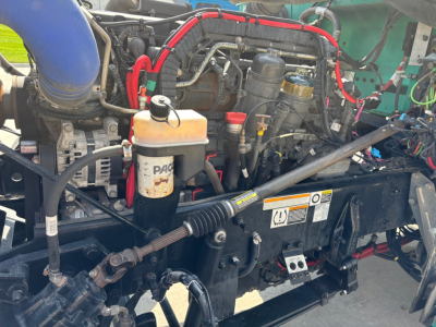 Peterbilt 389 engine bay showing coolant reservoir, hoses, and various components; visible lines, cables, and a yellowish coolant tank cap.