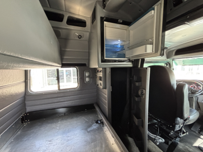 Peterbilt 389 truck interior showing cabin with open small refrigerator/freezer and overhead storage, front seats, and empty sleeper area.