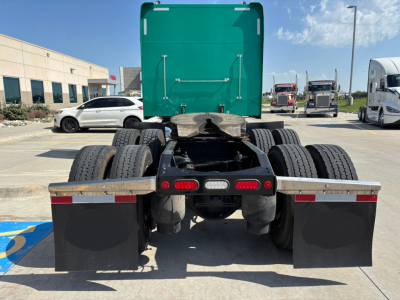 Peterbilt 389 tractor rear view showing dual rear axles, fifth wheel, rear mud flaps, and fuel/air lines. Green sleeper cab in distance, lot with other trucks.