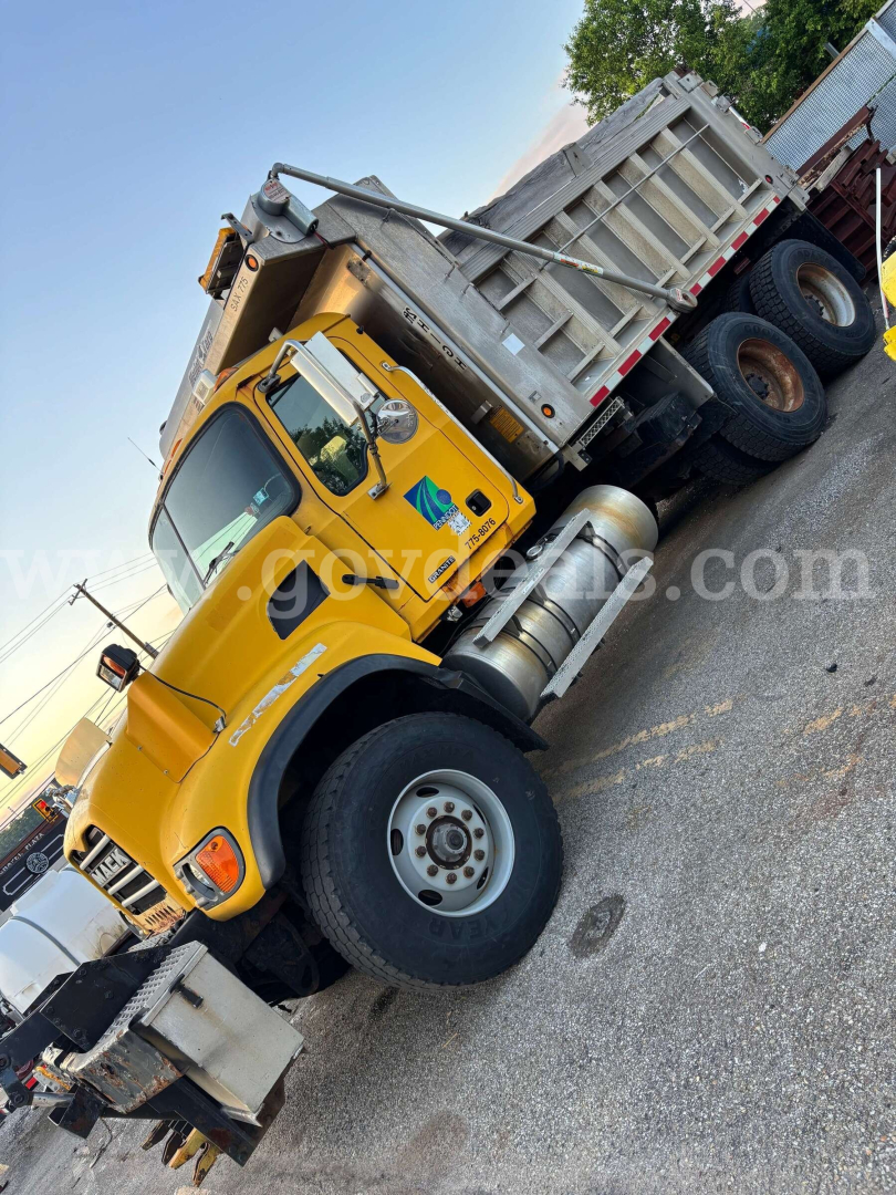 Mack CV713 dump truck shown in side angle with yellow cab and gray dump bed, front wheel visible.