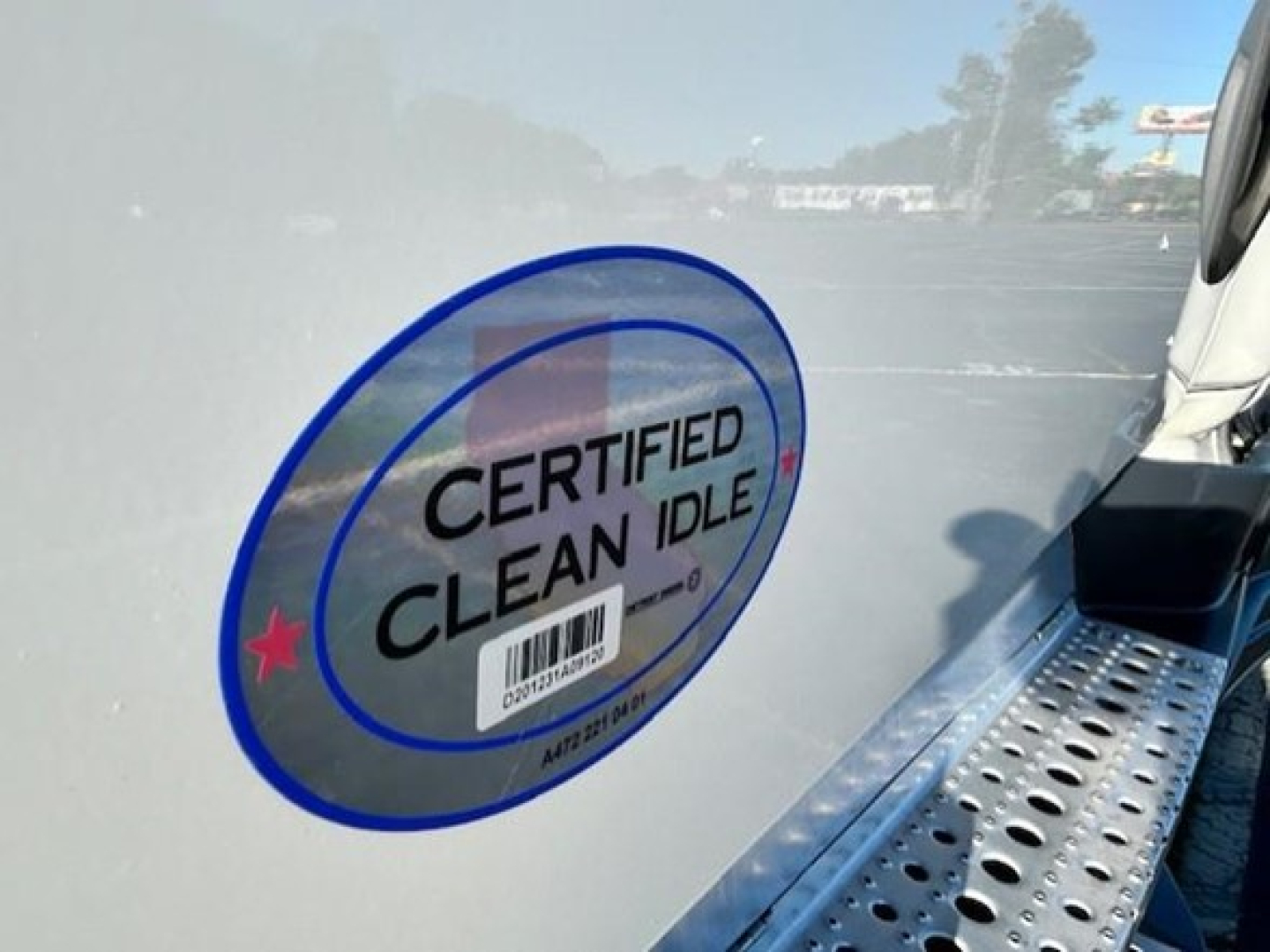 Sticker on Freightliner Cascadia showing CERTIFIED CLEAN IDLE and barcode on a light-colored truck door near a perforated step.