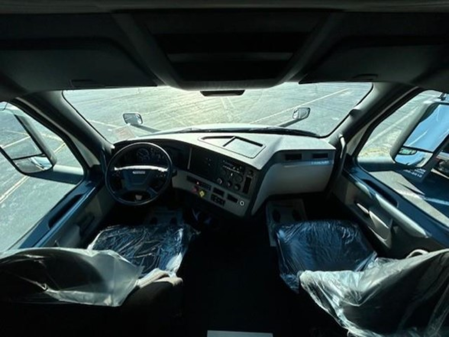 Cab interior of a Freightliner Cascadia 116 with plastic-covered seats, dashboard, steering wheel, and large windshield, showing driver and passenger view.