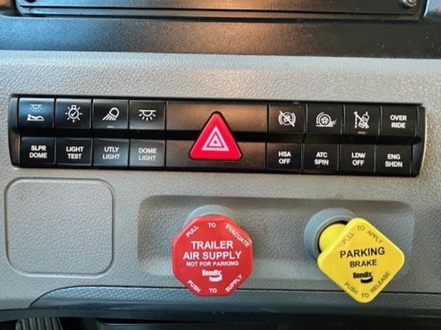 Truck dash panel showing hazard triangle button, various control icons, red trailer air supply cap, and yellow parking brake cap.