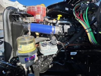 Truck engine bay with liquid-filled reservoirs and blue silicone hose; visible hydraulic reservoir, coolant cap, and electrical looms in a Freightliner Cascadia engine bay.