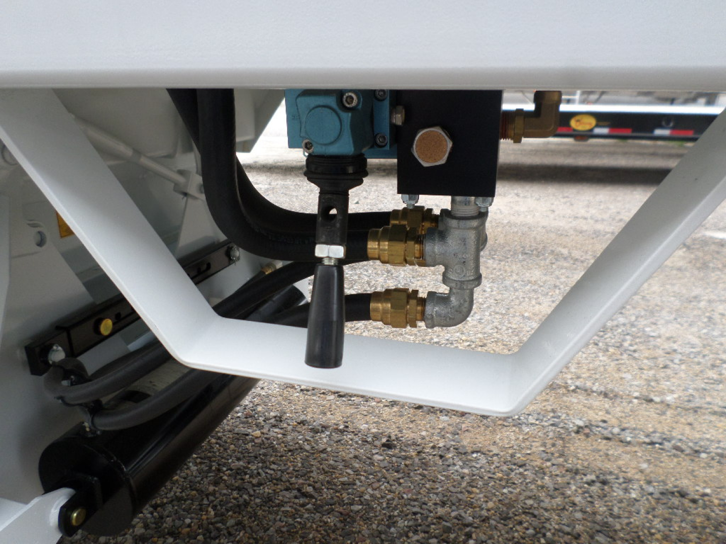 Trailer undercarriage with blue hydraulic valve and brass fittings connected to black hoses inside a white frame. Close-up view of plumbing and fittings on a new dump trailer.